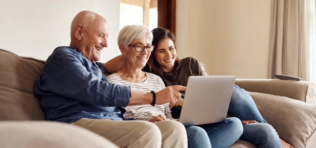 Baby Boomer couple sitting on a couch with an adult family member, smiling while reviewing information together on a laptop at home.