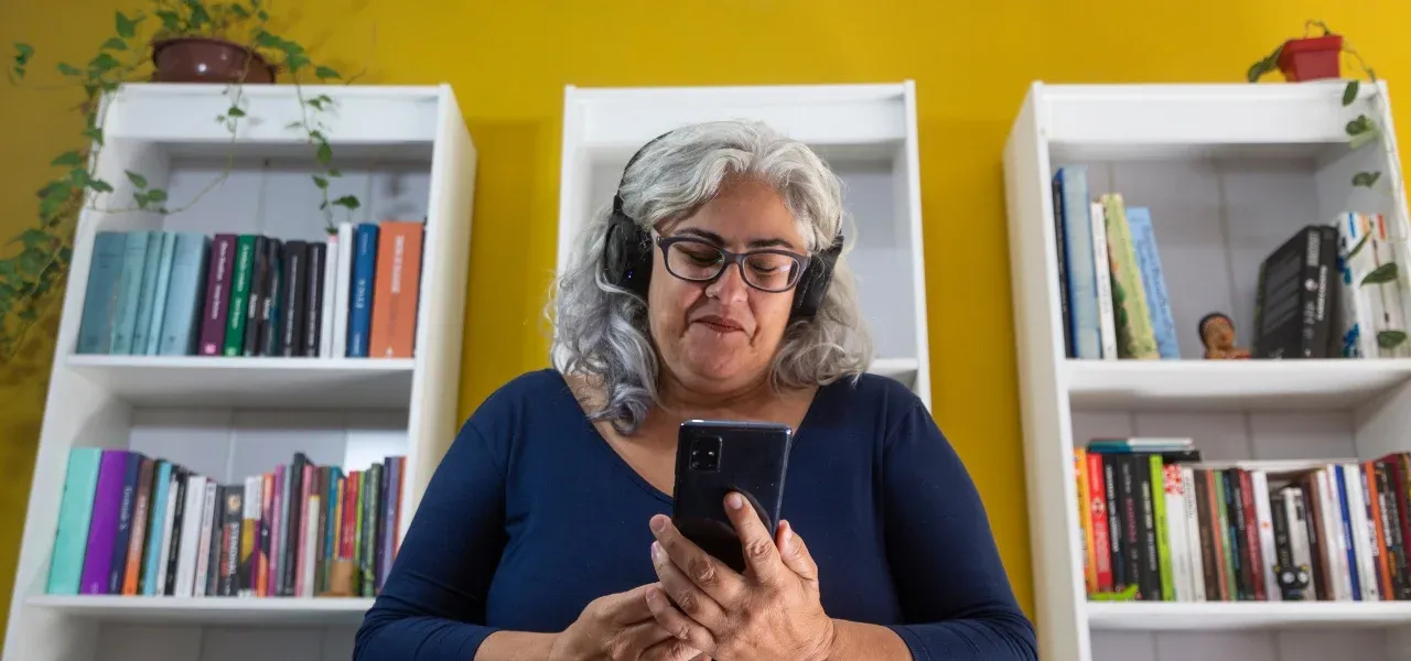 Gen X woman wearing headphones and glasses, standing in front of bookshelves while checking her smartphone at home.