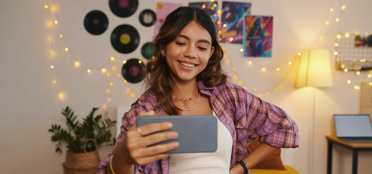 Gen Z woman smiling while watching a video on her smartphone in a cozy bedroom decorated with string lights and vinyl records.