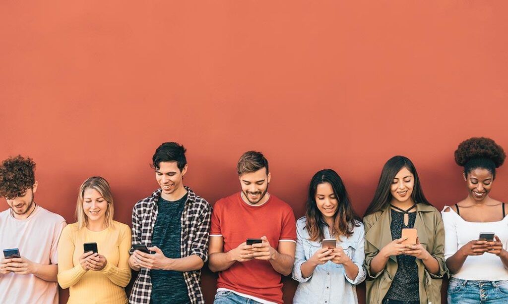 a line of young people leaning up against a red wall starring down at their phones