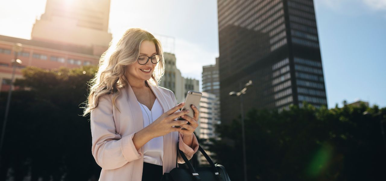 Professional woman standing outdoors in a city, smiling while using her smartphone.