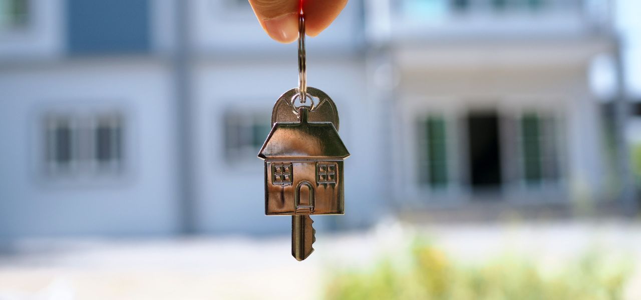 Hand holding a house key with a small house-shaped keychain in front of a blurred home exterior.